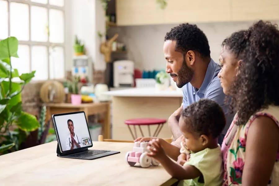 A young Black family with their toddler at the kitchen table, on a video call with a Black clinician shown on the screen